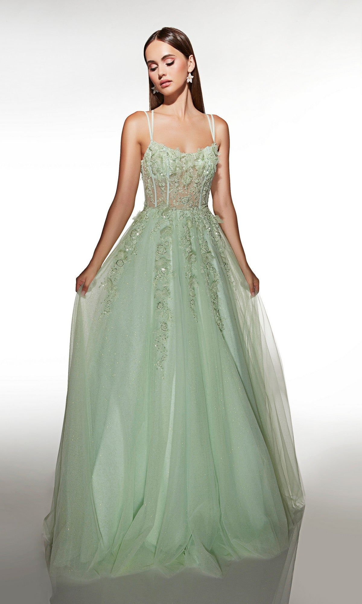 A woman wears the Alyce 62203 Long Prom Dress—a light green, sleeveless gown with 3D floral appliqué, sheer details, and a subtle corset bodice—standing against a plain white background.